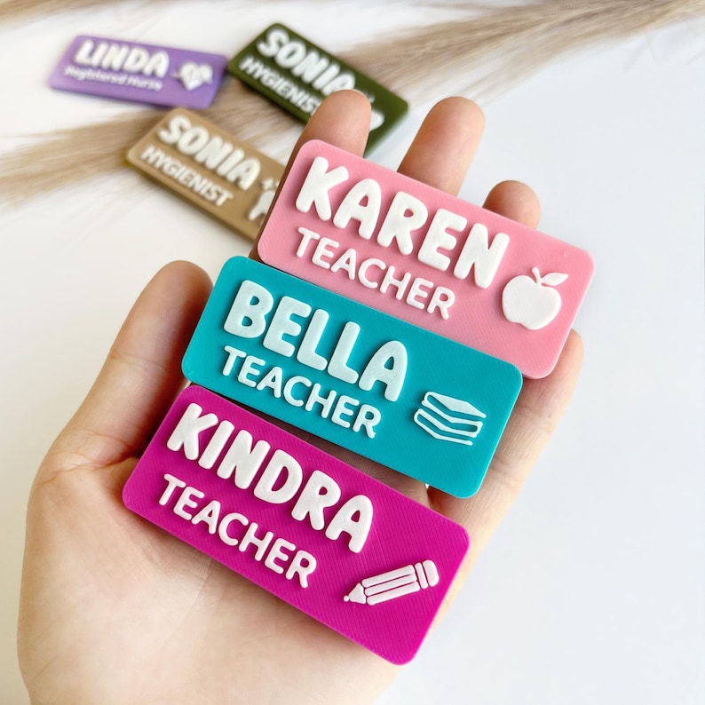 May include: Three rectangular name badges with raised white lettering. The top badge is pink with the text KAREN TEACHER and an apple icon. The middle badge is teal with BELLA TEACHER and a book icon. The bottom badge is magenta with KINDRA TEACHER and a pencil icon.
