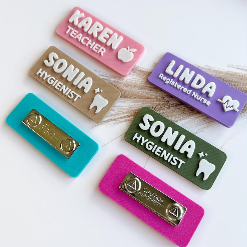 May include: A collection of rectangular name badges in pink, tan, purple, teal, olive green, and magenta. Each badge features raised white lettering and icons including an apple, tooth, and heart. Two badges show the silver magnetic backing.