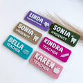 May include: Six rectangular name badges in purple, tan, olive green, teal, magenta, and pink. Each badge features raised white lettering and icons including a heart, tooth, book stack, pencil, and apple, displaying names and professional titles.