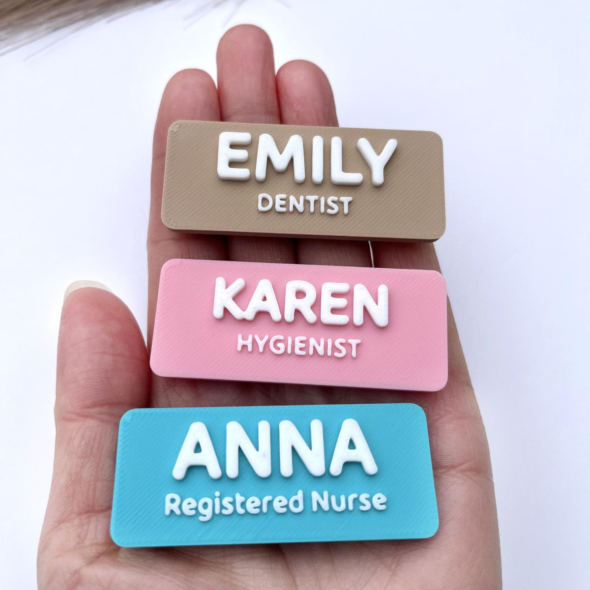 May include: Three rectangular name badges. The top badge is tan with the name "EMILY" and the title "DENTIST" in white. The middle badge is pink with "KAREN" and "HYGIENIST" in white. The bottom badge is blue with "ANNA" and "Registered Nurse" in white.