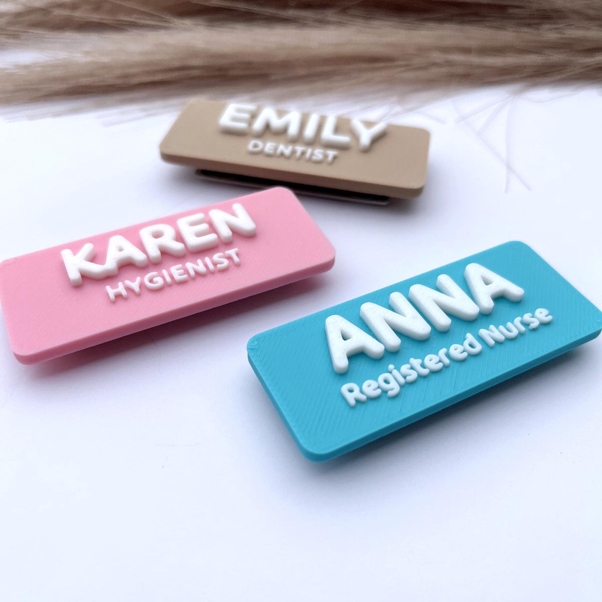 May include: Three rectangular name badges. One is beige with "EMILY" and "DENTIST" in white. Another is pink with "KAREN" and "HYGIENIST" in white. The last is turquoise with "ANNA" and "Registered Nurse" in white.