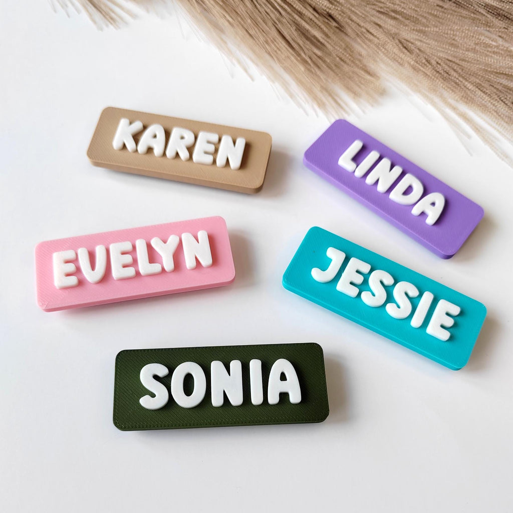 May include: Five rectangular name tags in various colours: tan, purple, pink, teal, and olive green. Each tag features a different name in white, raised lettering: Karen, Linda, Evelyn, Jessie, and Sonia. The tags are arranged on a white surface.
