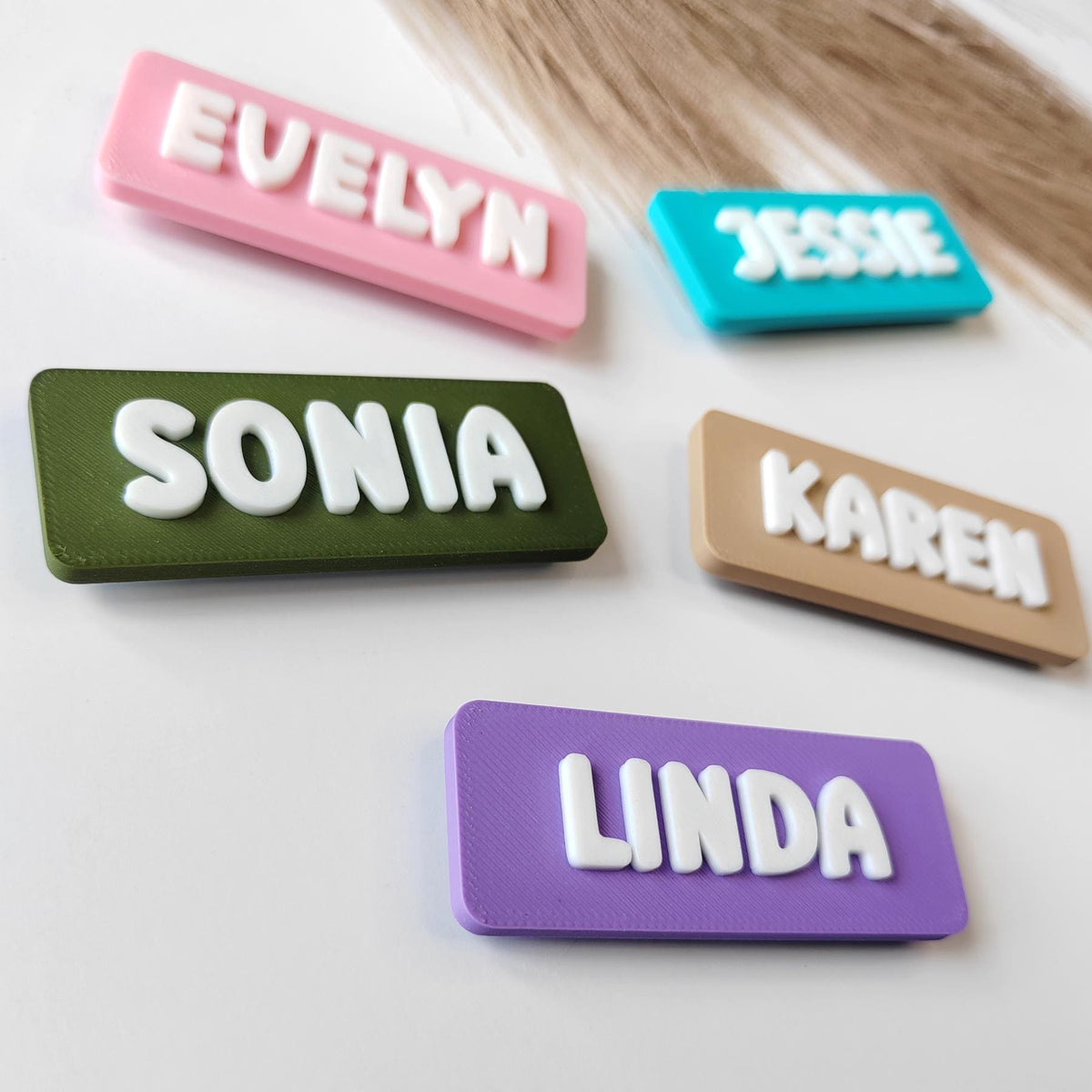 May include: A selection of name tags in various colours, each with a different name in white, raised lettering. The rectangular tags are in shades of pink, teal, green, tan, and purple. The names displayed are Evelyn, Jessie, Sonia, Karen, and Linda.