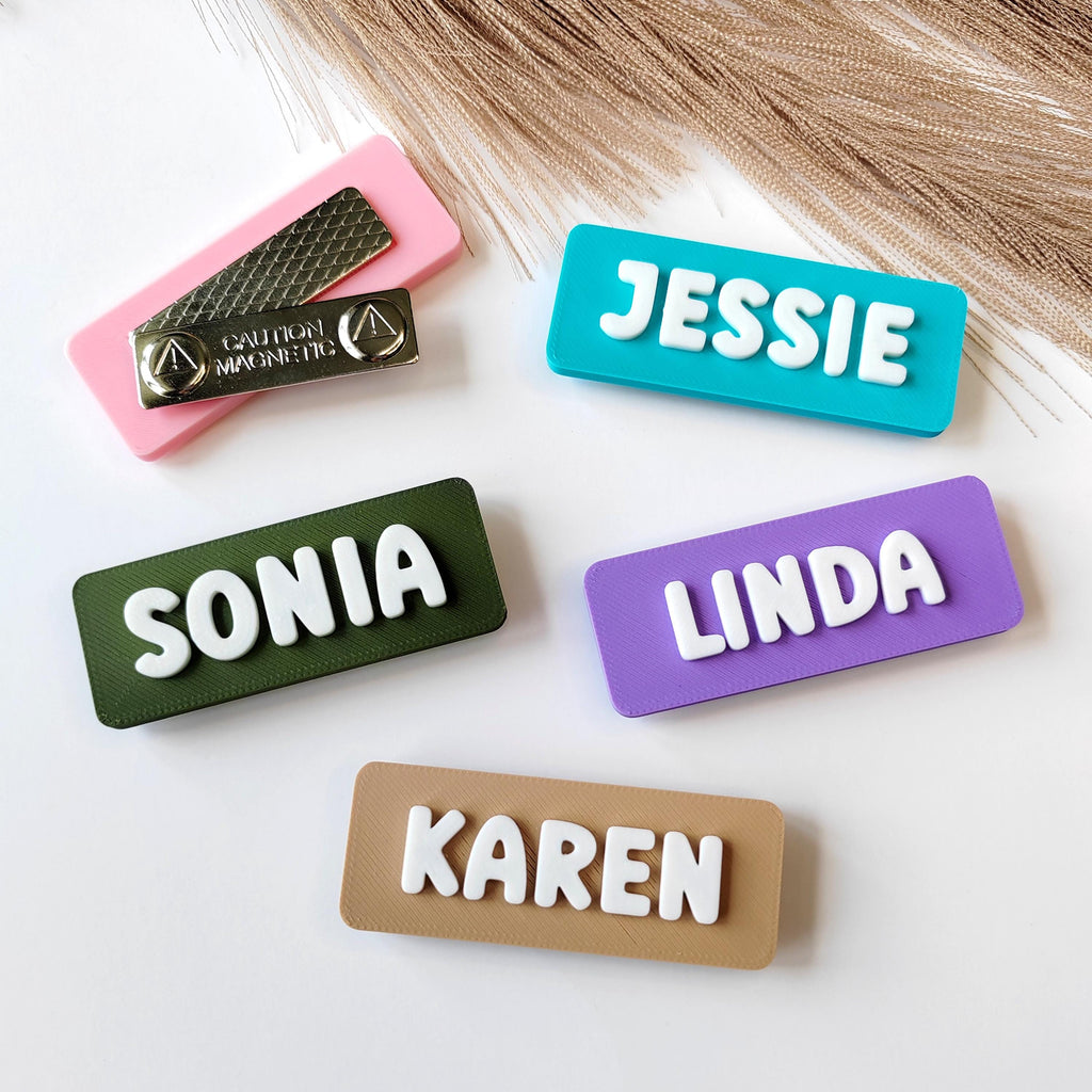 May include: A selection of colourful name tags with white raised lettering. The tags are in various colours, including teal, purple, green, and tan. The names "Jessie", "Linda", "Sonia", and "Karen" are displayed. A pink tag with a magnetic backing is also present.