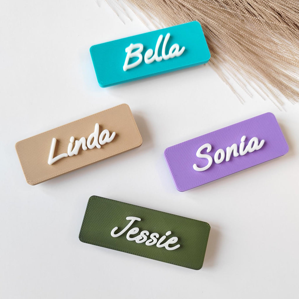 May include: Four rectangular name tags in teal, beige, lilac, and olive green. Each tag has a different name in white cursive lettering: Bella, Linda, Sonia, and Jessie. The tags are arranged on a white surface.