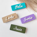 May include: Four rectangular name tags in teal, beige, lilac, and olive green. Each tag has a different name in white cursive lettering: Bella, Linda, Sonia, and Jessie. The tags are arranged on a white surface.