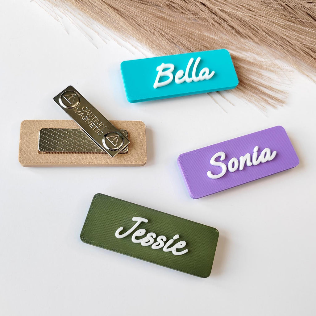 May include: Four name tags in teal, purple, and green with white 3D lettering. One tag has a magnetic backing with the text "CAUTION MAGNETIC". The names "Bella", "Sonia", and "Jessie" are displayed.