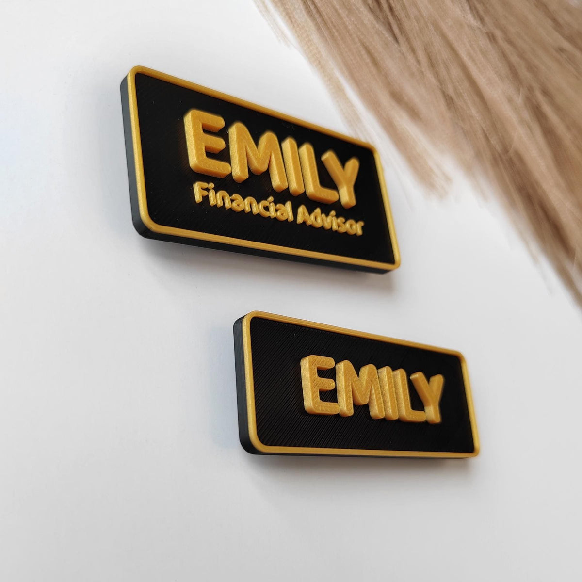 May include: Two black and gold name badges. The top badge reads "EMILY Financial Advisor" in gold lettering. The bottom badge reads "EMILY" in gold lettering. The badges are rectangular with rounded corners.
