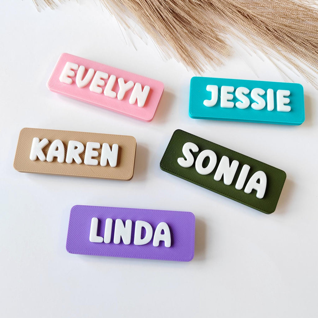 May include: A selection of personalised name tags in shades of pink, teal, beige, green, and purple. Each tag displays a different name in white, raised lettering: Evelyn, Jessie, Karen, Sonia, and Linda. The tags are rectangular with a textured surface.