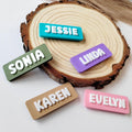 May include: A selection of colourful name tags with white raised lettering. The tags are rectangular and feature the names "Jessie", "Sonia", "Linda", "Karen", and "Evelyn". The tags are in various colours, including teal, green, purple, tan, and pink.