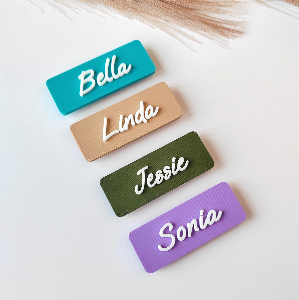 May include: Four rectangular name tags in teal, tan, olive green, and purple. Each tag features a white cursive name: Bella, Linda, Jessie, and Sonia. The tags are arranged on a white surface, with a few strands of dried grass in the background.