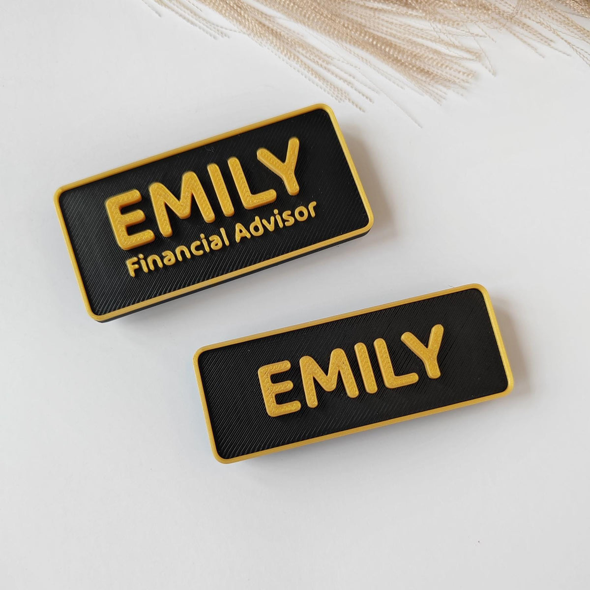 May include: Two black and gold name badges. The name "EMILY" is printed in gold on each badge. One badge also includes the title "Financial Advisor" in gold. The badges have a textured surface and a gold border.